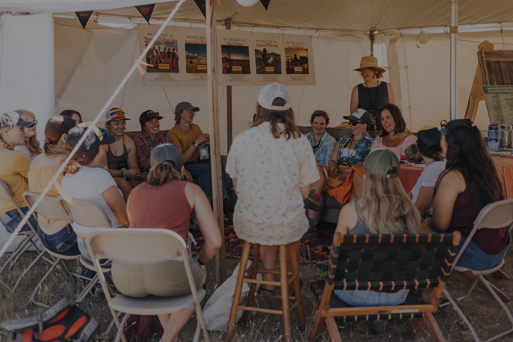 A group of people sit in a circle under a tent, listening and chatting during a casual gathering or discuss