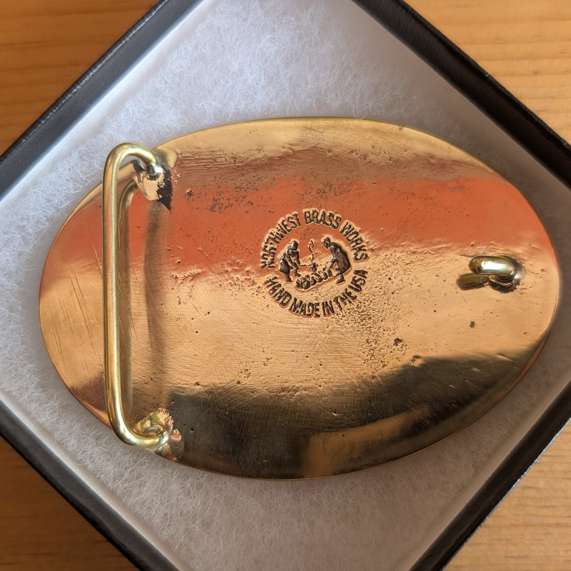 The back of a brass belt buckle with makers engraved design on a wooden surface