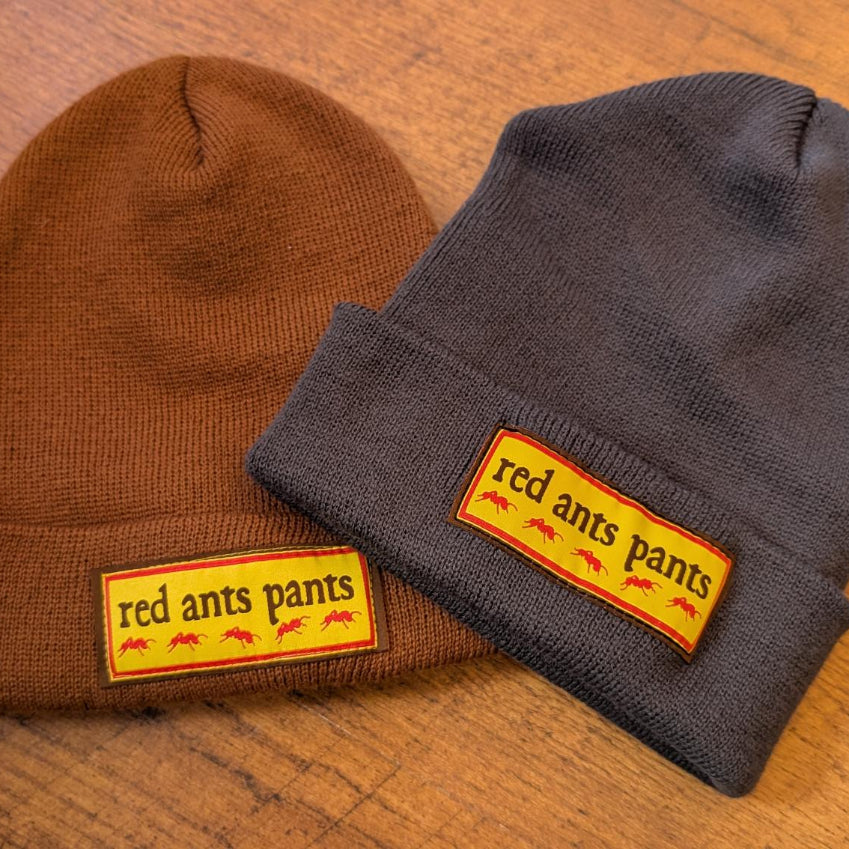 Two knit beanie hats on a wooden surface: one brown and one dark gray, each with a folded cuff and a rectangular yellow patch reading 'red ants pants' with small red ant illustrations.