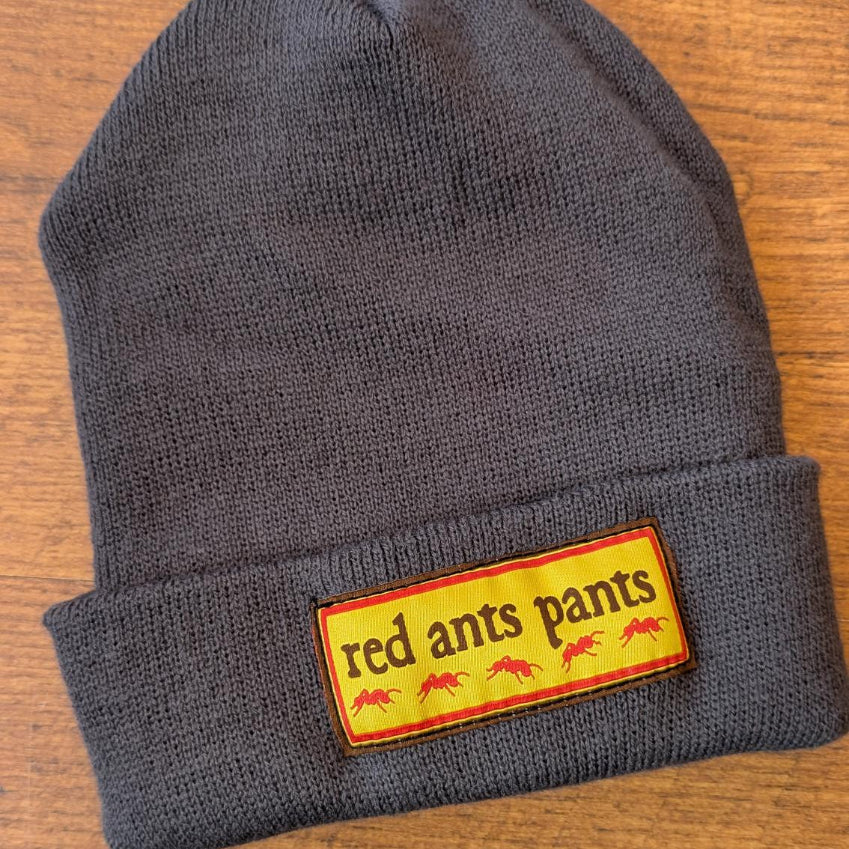 A dark gray knit beanie with a folded cuff, featuring a rectangular yellow patch on the front with the text 'red ants pants' and small red ant illustrations, placed on a wooden surface. - Iron Gray